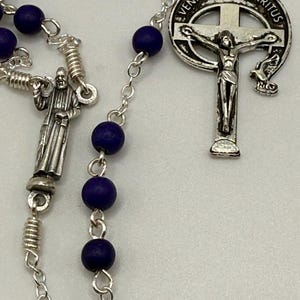 May include: A rosary with dark blue beads and silver-toned metal accents. The rosary features a crucifix pendant with the inscription "VENI CREATOR SPIRITUS" and a figure of Jesus Christ. A small figure of a saint is also present.
