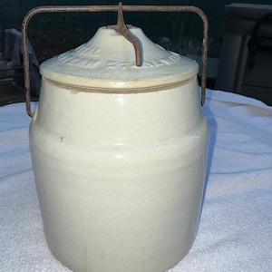 Pode incluir: Pote de grés branco sujo com tampa e alça de metal enferrujado. A tampa tem um padrão decorativo em relevo. O pote é cilíndrico com um corpo ligeiramente cónico. Um recipiente de cozinha vintage.