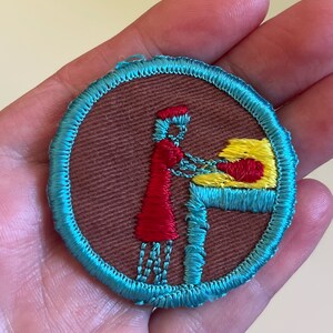 May include: A round, blue-trimmed patch with a brown background. A red-dressed figure with a yellow hat is shown standing at a table with a yellow object on it.