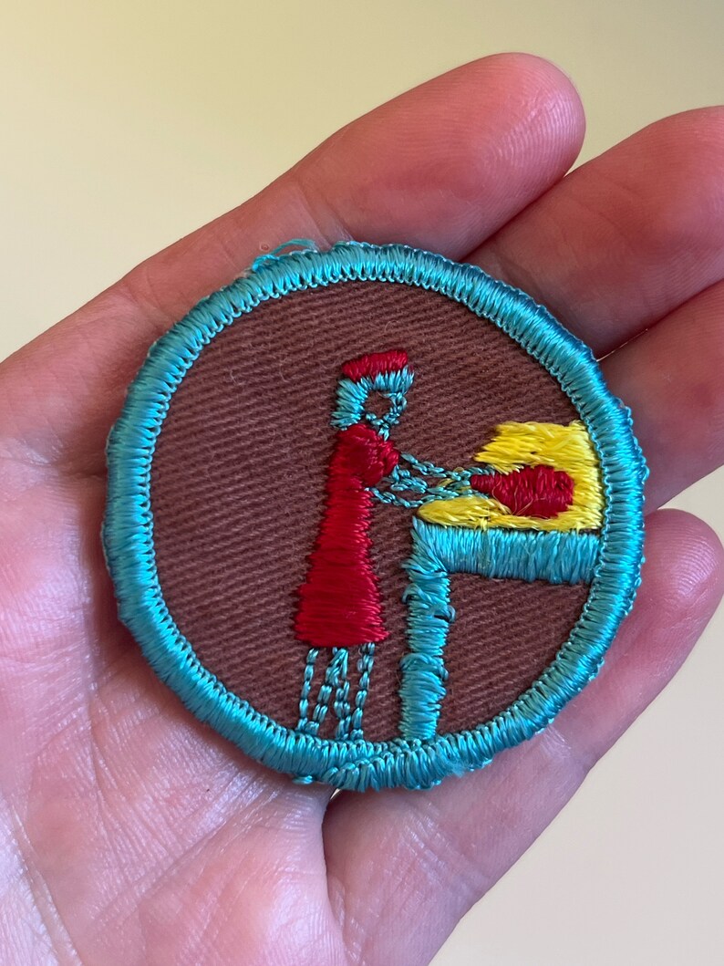 May include: A round, blue-trimmed patch with a brown background. A red-dressed figure with a yellow hat is shown standing at a table with a yellow object on it.