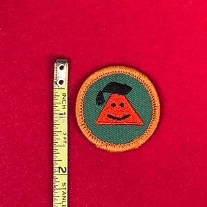 May include: A green and orange embroidered scout patch with a smiling red triangle wearing a black graduation cap.