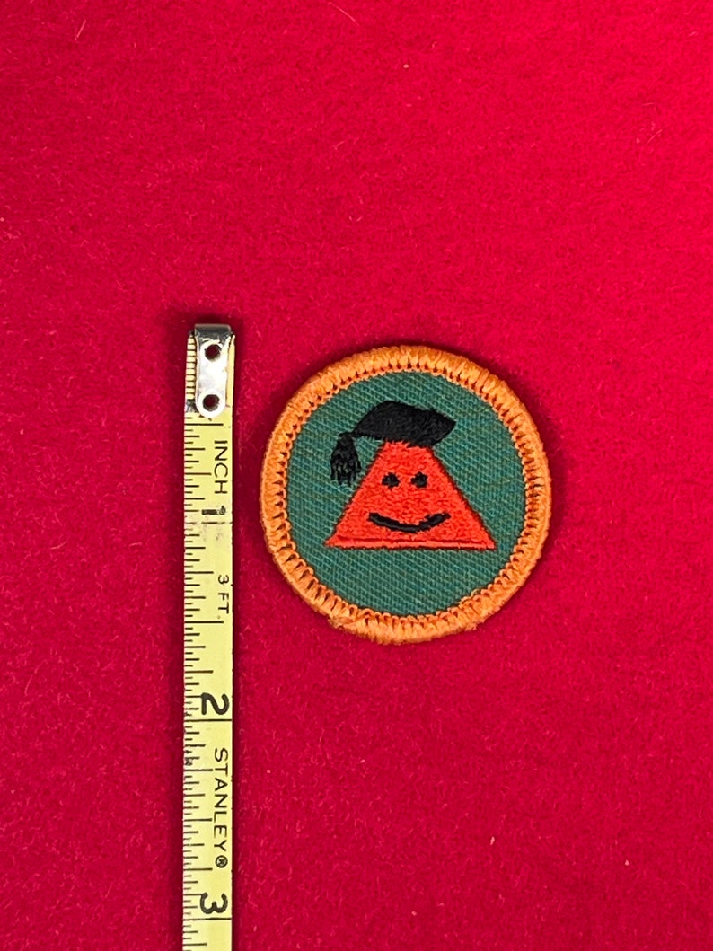 May include: A green and orange embroidered scout patch with a smiling red triangle wearing a black graduation cap.