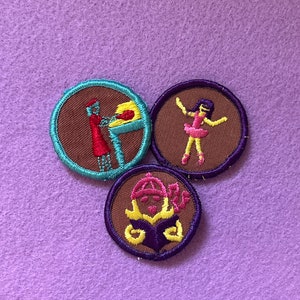 May include: Three embroidered Girl Scout badges with brown felt centers and colorful embroidered borders. The badges depict a girl baking, a girl dancing, and a girl reading a book.