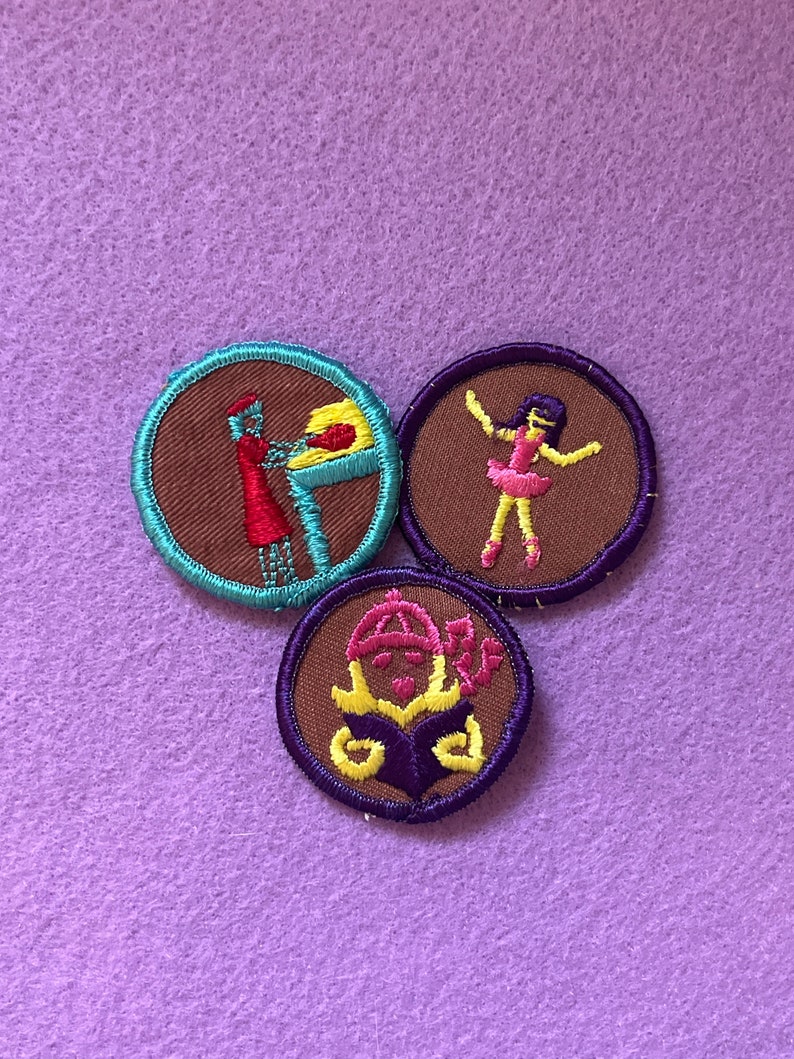 May include: Three embroidered Girl Scout badges with brown felt centers and colorful embroidered borders. The badges depict a girl baking, a girl dancing, and a girl reading a book.
