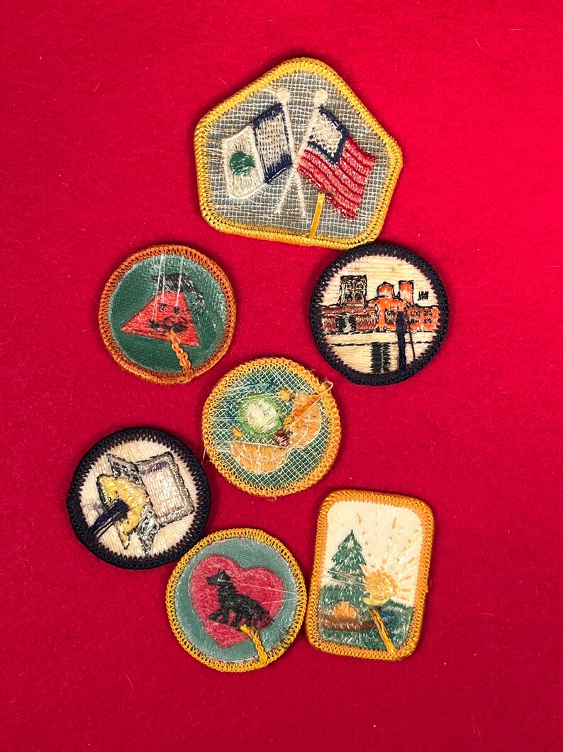 May include: Seven embroidered patches with yellow stitching. The patches feature various designs, including flags, a building, a tree, a wolf, and a campfire.