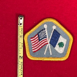 May include: A blue and gold embroidered patch with two crossed flags. The flags are the American flag and a white flag with a green shamrock. The patch is about 2 inches wide.