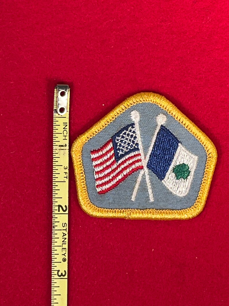 May include: A blue and gold embroidered patch with two crossed flags. The flags are the American flag and a white flag with a green shamrock. The patch is about 2 inches wide.