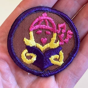 May include: A brown embroidered patch with a purple and yellow design. The design features a person wearing a pink hat and holding a book.