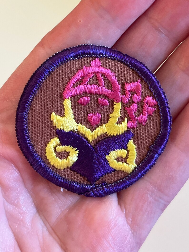 May include: A brown embroidered patch with a purple and yellow design. The design features a person wearing a pink hat and holding a book.