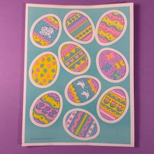May include: A sheet of Easter egg stickers with various designs. The eggs are decorated with patterns like polka dots, stripes, and floral motifs, and some feature images of a sheep, bunnies, and butterflies. The eggs are outlined in white and set against a light blue background.