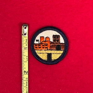 May include: A blue and yellow embroidered patch with a depiction of a factory building. The patch is circular and has a dark blue border.