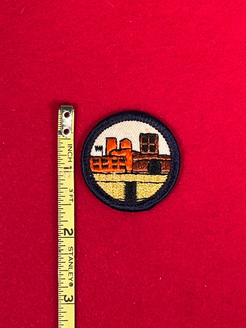 May include: A blue and yellow embroidered patch with a depiction of a factory building. The patch is circular and has a dark blue border.