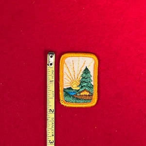 May include: A yellow and green embroidered patch with a sunrise, a pine tree, and a tent. The patch has a yellow border and white stitching.
