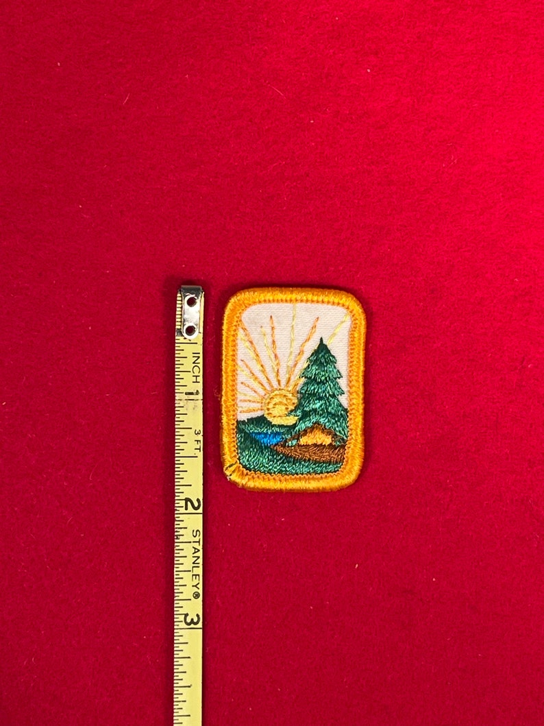 May include: A yellow and green embroidered patch with a sunrise, a pine tree, and a tent. The patch has a yellow border and white stitching.