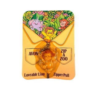 May include: An orange lion-shaped zipper pull attached to a yellow cardboard backing. The packaging features illustrations of a purple elephant, a monkey, and a lion in a garden. Text on the card reads AVON, ZIP A ZOO, and Loveable Lion Zipper Pull.