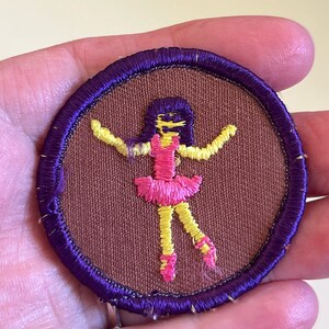 May include: A purple and brown embroidered patch featuring a yellow and pink stick figure ballerina with black hair.