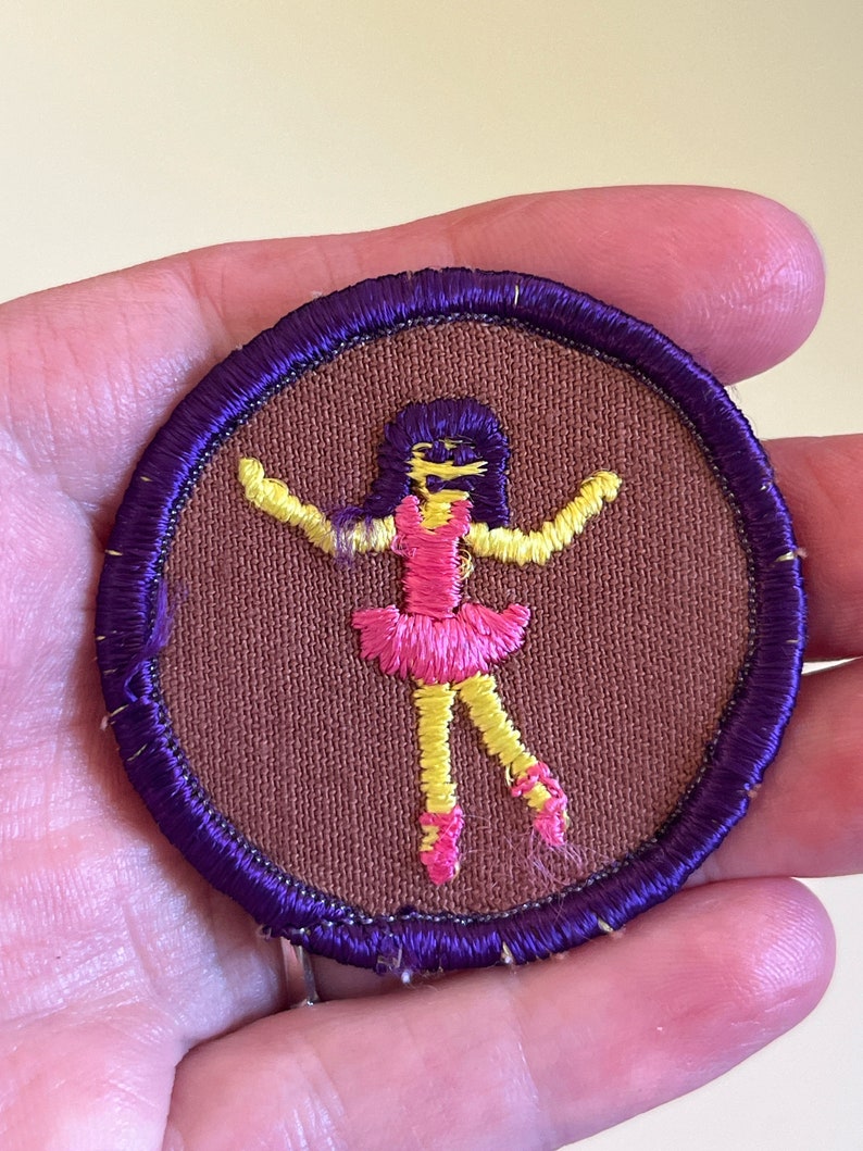 May include: A purple and brown embroidered patch featuring a yellow and pink stick figure ballerina with black hair.
