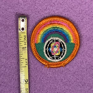Puede incluir: Un parche redondo bordado con un arco&iacute;ris, un globo terr&aacute;queo y el texto "World" en un c&iacute;rculo negro. El parche est&aacute; rodeado por un borde verde y un borde naranja.