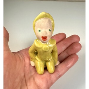 May include: A small, vintage ceramic figurine of a seated figure in a yellow jumpsuit and hood. The face is pale with green eyes and a red mouth. The figurine is held in a hand against a white background.