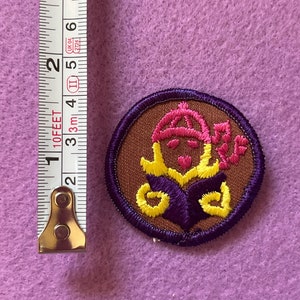 May include: A brown embroidered patch with a purple border. The patch features a yellow and pink design of a person wearing a hat and holding a flower.