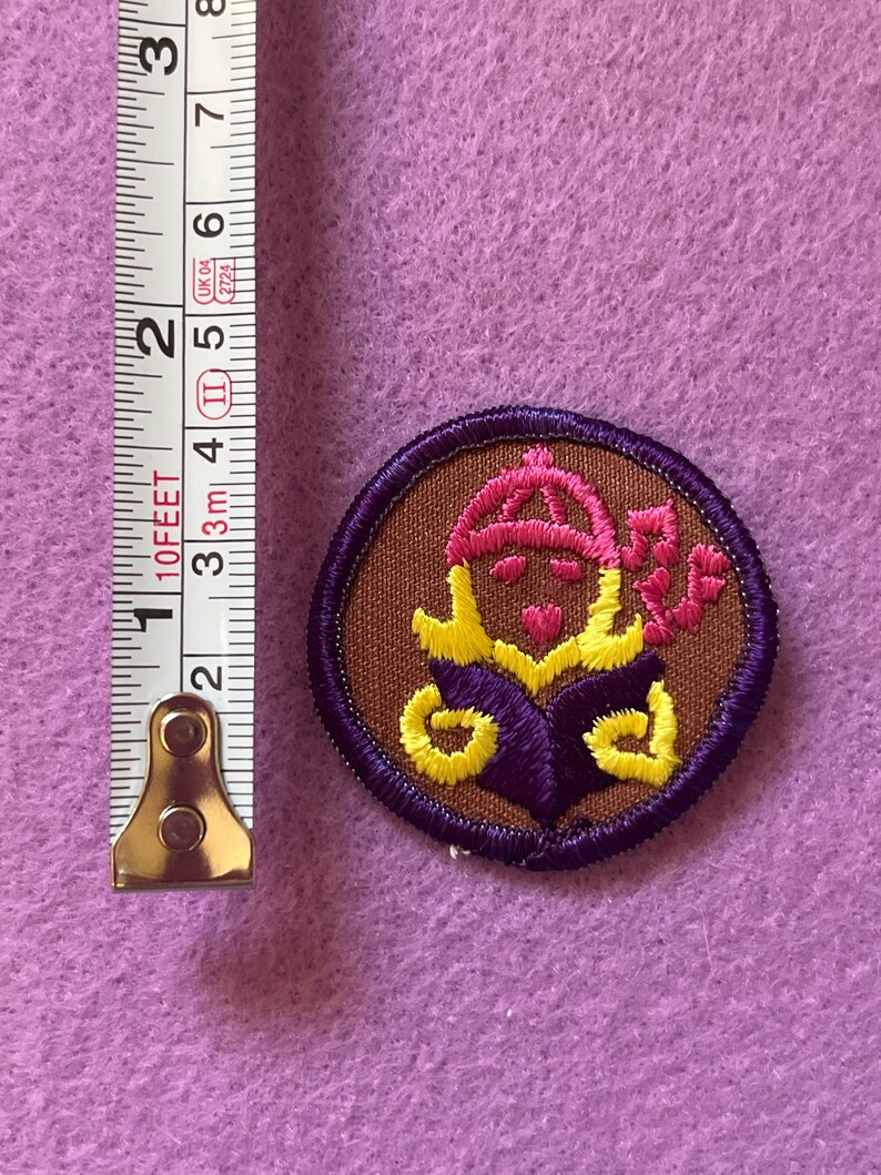 May include: A brown embroidered patch with a purple border. The patch features a yellow and pink design of a person wearing a hat and holding a flower.