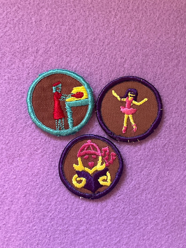 May include: Three embroidered Girl Scout badges on a purple background. The badges are circular and feature a brown background with colorful embroidery. The first badge shows a person in a red dress making a loaf of bread. The second badge shows a person in a pink dress dancing. The third badge shows a person in a yellow hat reading a book.