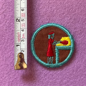 May include: A brown and teal embroidered patch with a woman in a red dress and a yellow hat standing at a table with a yellow loaf of bread. The patch is about 1 inch in diameter.
