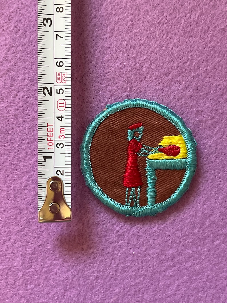 May include: A brown and teal embroidered patch with a woman in a red dress and a yellow hat standing at a table with a yellow loaf of bread. The patch is about 1 inch in diameter.