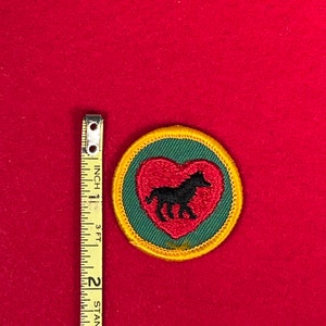 May include: A green and red embroidered patch with a black horse inside a heart. The patch is surrounded by a yellow border.