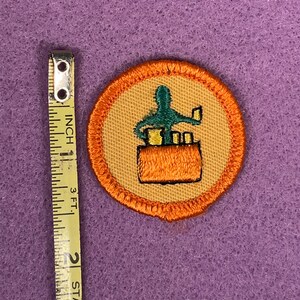 Puede incluir: Una insignia de Girl Scout redonda, con borde naranja, de color beige con una figura verde que sostiene un libro y una pila de libros en un bolsillo marr&oacute;n. El texto "Bookworm" est&aacute; impreso en la insignia.