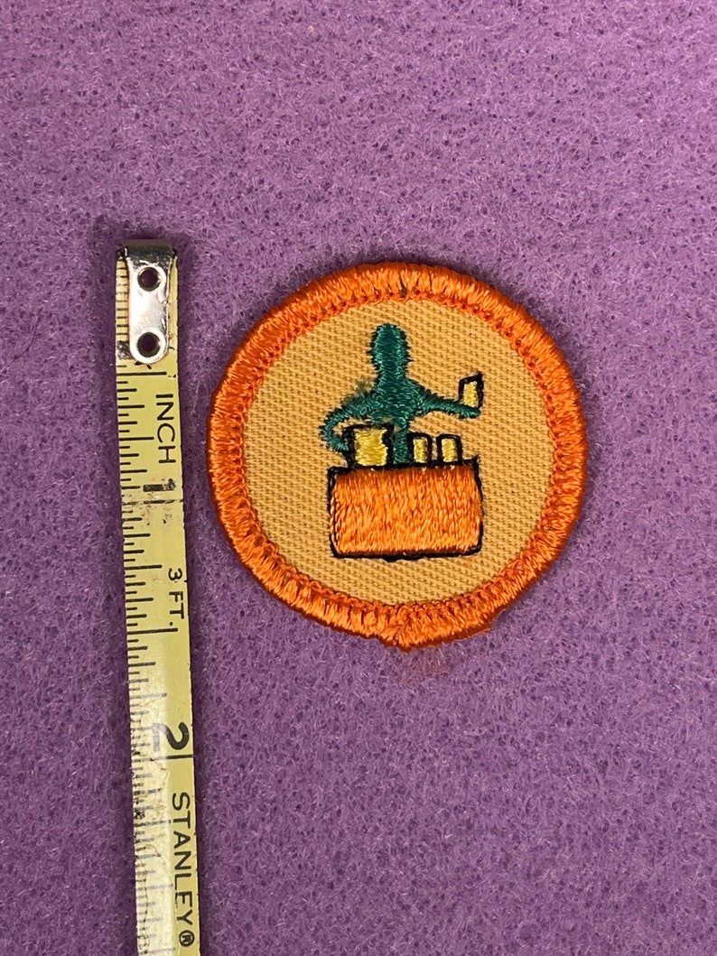 Puede incluir: Una insignia de Girl Scout redonda, con borde naranja, de color beige con una figura verde que sostiene un libro y una pila de libros en un bolsillo marr&oacute;n. El texto "Bookworm" est&aacute; impreso en la insignia.