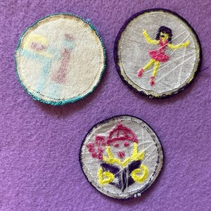 May include: Three embroidered iron-on patches with white fabric backgrounds. The first patch is teal and yellow with a cartoon of a person holding a paintbrush. The second patch is purple and yellow with a cartoon of a person in a pink dress dancing. The third patch is purple and yellow with a cartoon of a person wearing a hat and holding a book.