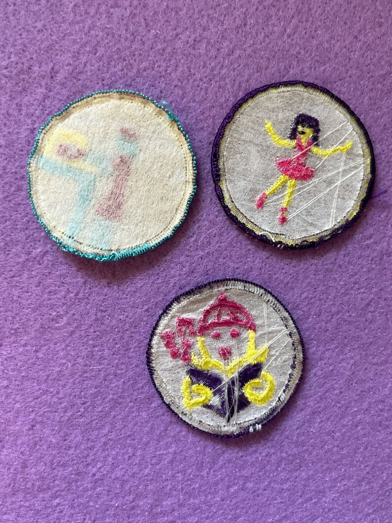 May include: Three embroidered iron-on patches with white fabric backgrounds. The first patch is teal and yellow with a cartoon of a person holding a paintbrush. The second patch is purple and yellow with a cartoon of a person in a pink dress dancing. The third patch is purple and yellow with a cartoon of a person wearing a hat and holding a book.