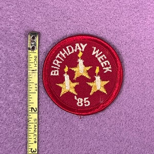 Puede incluir: Parche de fieltro rojo de la semana de cumplea&ntilde;os con tres estrellas doradas y velas blancas. El texto "Birthday Week" est&aacute; en blanco y el a&ntilde;o "'85" est&aacute; en blanco.