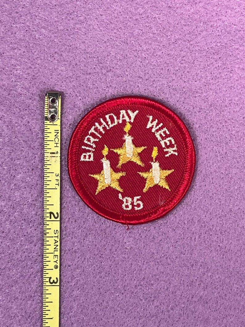 Puede incluir: Parche de fieltro rojo de la semana de cumplea&ntilde;os con tres estrellas doradas y velas blancas. El texto "Birthday Week" est&aacute; en blanco y el a&ntilde;o "'85" est&aacute; en blanco.