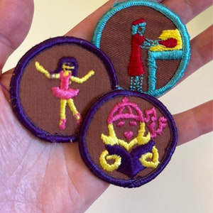 May include: Three embroidered patches with brown felt backgrounds and colorful stitching. One patch features a girl in a pink dress and yellow shoes, another shows a person in a red dress baking, and the third shows a girl with a crown and a purple book.