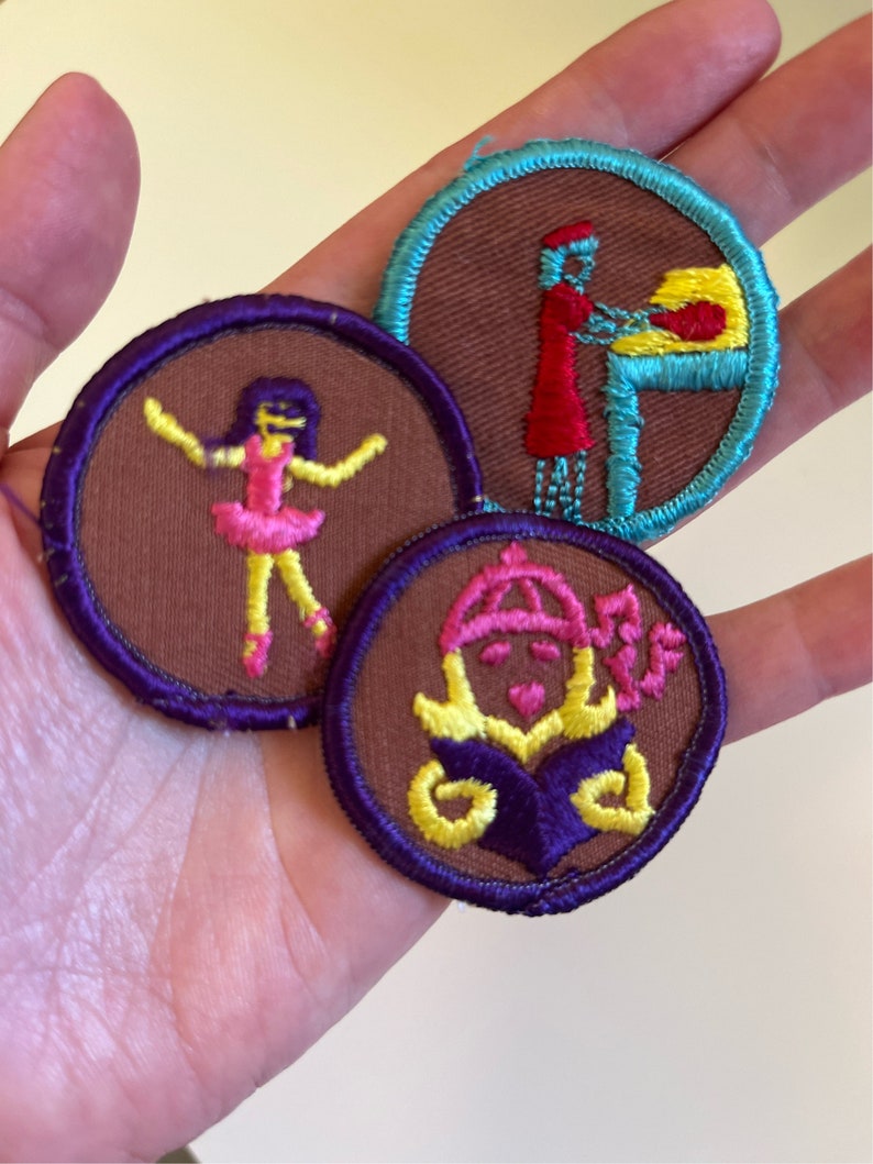 May include: Three embroidered patches with brown felt backgrounds and colorful stitching. One patch features a girl in a pink dress and yellow shoes, another shows a person in a red dress baking, and the third shows a girl with a crown and a purple book.
