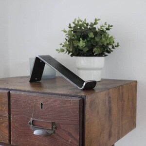 May include: A black metal shelf bracket with a silver finish, mounted on a wooden dresser with a drawer. A small potted plant sits on the dresser.