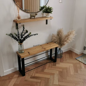 Puede incluir: Una estanter&iacute;a de madera con soportes de metal negro, un peque&ntilde;o banco de madera con patas de metal negro y un zapatero de metal negro, y una figurita de animal de madera decorativa. La estanter&iacute;a y el banco est&aacute;n contra una pared blanca con un espejo redondo encima de la estanter&iacute;a. Hay plantas en macetas en la estanter&iacute;a y un jarr&oacute;n con flores secas en el suelo.