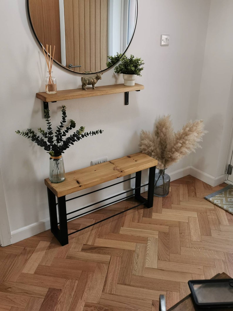 Puede incluir: Una estanter&iacute;a de madera con soportes de metal negro, un peque&ntilde;o banco de madera con patas de metal negro y un zapatero de metal negro, y una figurita de animal de madera decorativa. La estanter&iacute;a y el banco est&aacute;n contra una pared blanca con un espejo redondo encima de la estanter&iacute;a. Hay plantas en macetas en la estanter&iacute;a y un jarr&oacute;n con flores secas en el suelo.