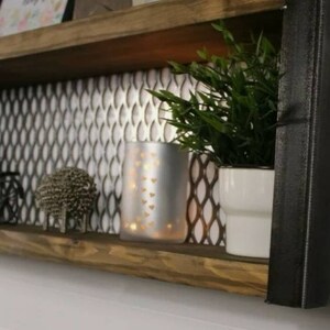 May include: A wooden shelf with two tiers, featuring a black metal frame with a diamond pattern. The shelf is decorated with a silver metallic tile backsplash and has various decorative items, including a blue ceramic vase, a silver candle holder, and a small metal sculpture.
