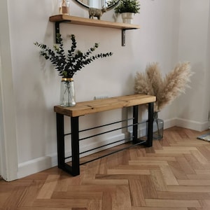 Puede incluir: Un banco de madera con un marco de metal negro y tres estantes para guardar zapatos. El banco est&aacute; en un pasillo con un suelo de madera en espiga.