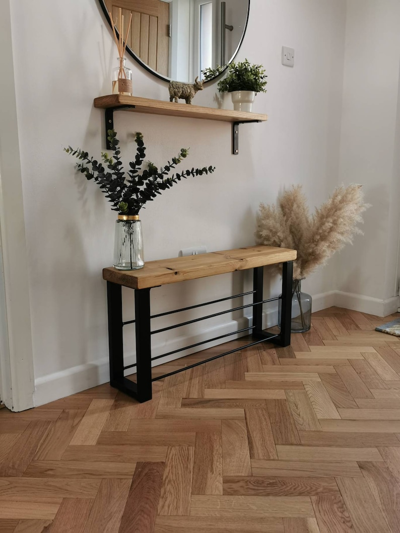 Puede incluir: Un banco de madera con un marco de metal negro y tres estantes para guardar zapatos. El banco est&aacute; en un pasillo con un suelo de madera en espiga.