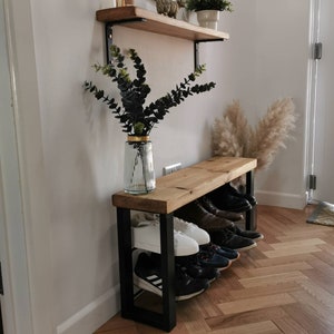 Puede incluir: Un banco de madera con un marco de metal negro y dos estantes para guardar zapatos. El banco est&aacute; en un pasillo con suelo de madera y una pared blanca. Hay plantas y una estanter&iacute;a en la pared sobre el banco.