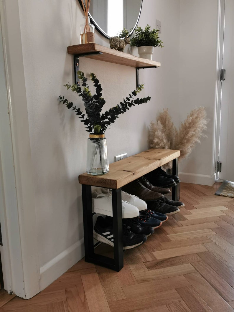 Puede incluir: Un banco de madera con un marco de metal negro y dos estantes para guardar zapatos. El banco est&aacute; en un pasillo con suelo de madera y una pared blanca. Hay plantas y una estanter&iacute;a en la pared sobre el banco.