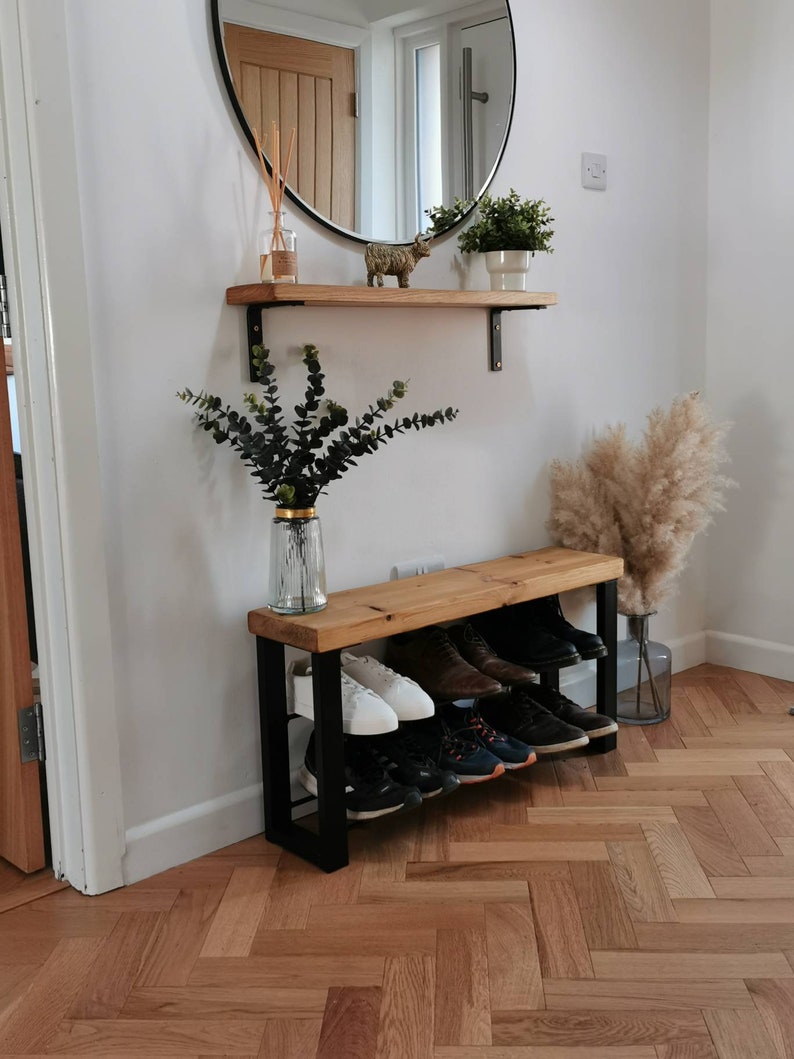 Puede incluir: Un banco de madera con un marco de metal negro y dos estantes para guardar zapatos. El banco est&aacute; en un pasillo con una pared blanca y un espejo redondo encima.