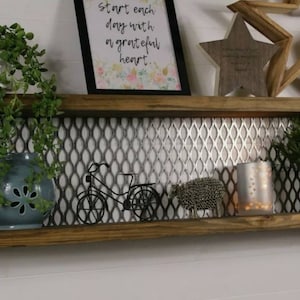 May include: A rustic wooden shelf with metal accents features a silver mesh backing. The shelf is decorated with a framed print that reads "Start each day with a grateful heart."  There are also two wooden stars, a small metal bicycle, a ceramic pot with a green plant, a silver candle holder, and a small ceramic pot with a green plant.