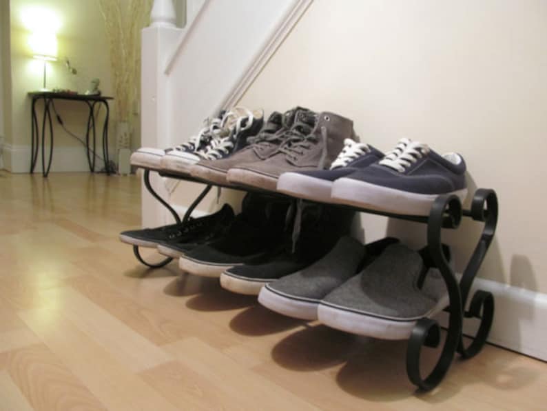 May include: A black metal shoe rack with two tiers holds a variety of trainers. The rack is in a hallway with light wood floors. The trainers are a mix of black, grey, and white.