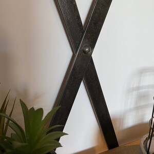 May include: Close-up of a black metal X-shaped shelf bracket with a distressed finish. The bracket is attached to a white wall and has a small metal rivet at the intersection of the two pieces.