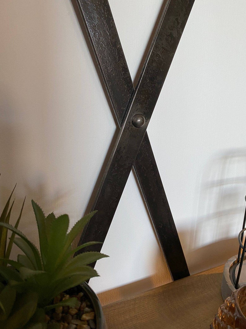 May include: Close-up of a black metal X-shaped shelf bracket with a distressed finish. The bracket is attached to a white wall and has a small metal rivet at the intersection of the two pieces.